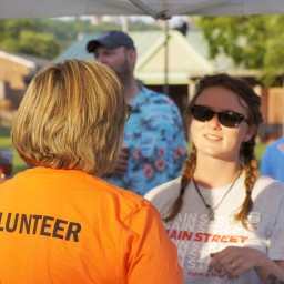 Volunteer in an orange shirt speaks with a Main Street staff member during an outdoor event, with others gathered casually in the background.