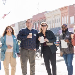 Six adults smile and hold coffee, pastries, and notebooks while standing on a sunny Main Street sidewalk lined with historic buildings and lamppost banners that read "Nevada Cubs."