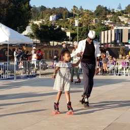 In East Oakland, California, a young girl and an older man hold hands while roller skating together at an outdoor community event hosted by the Black Cultural Zone.