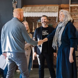 A group of five people greet each other inside a warmly lit coffee shop in Nevada, Iowa, with two individuals in the foreground shaking hands and others smiling in the background.