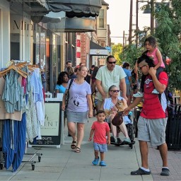 Shoppers stroll down a busy downtown sidewalk past a rolling rack of dresses and tops displayed outside a boutique. A child in a red shirt walks ahead of an adult pushing a stroller, while another adult carries a small child on their shoulders. Nearby, a person in a mobility scooter holds a small dog, and a street vendor’s jewelry stand and a yellow food truck peek out behind leafy trees.