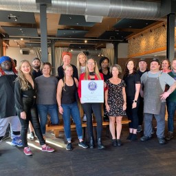 A group of about fifteen people stands together inside a modern restaurant space with wooden floors, high ceilings, and exposed ductwork. They wear a mix of work aprons, casual clothes, and business-casual outfits. In the center, one person holds a “Shop Small” sign that reads “AT Town Hall and The Dispensary.” Behind them are seating benches, tables, and an open kitchen area with tile accents on the walls.