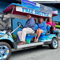 A custom-painted, tropical-themed golf cart parked on the street in front of a bright blue storefront, with a “FREE SHUTTLE” sign and sponsor banners overhead. Four passengers sit onboard—one at the wheel wearing a plaid shirt and light pants, two seated behind on blue cushions (one in a bright blue patterned dress), and one in a pink top—smiling at the camera. The cart’s body features palm tree and beach graphics, and its oversized off-road wheels are painted blue and white.