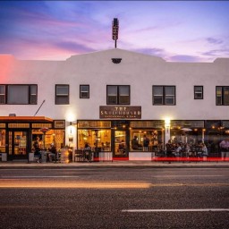 A two-story white stucco building at dusk, its ground floor glowing with warm light from large windows and outdoor seating areas on either side of a central entrance. A bronze sign above the door reads “The Switchboard Restaurant & Bar.” To the left, a neon vertical “Hotel” sign casts a red glow on the upper façade, while string lights and patio heaters illuminate diners outside. Faint streaks of red tail lights blur across the street in front, and a purple-pink sky fades into deep blue above.