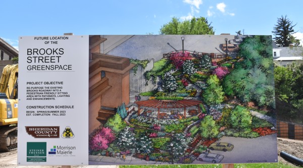 A construction site sign showing the future location of the Brooks Street Greenspace in Sheridan, Wyoming. The sign includes a colorful illustrated rendering of the planned park, featuring pathways, lush landscaping, a pergola seating area, and lighting. Text on the sign outlines the project objective and construction schedule, with logos from local partners and planning organizations.