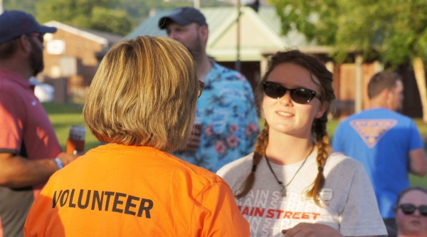 Volunteer in an orange shirt speaks with a Main Street staff member during an outdoor event, with others gathered casually in the background.