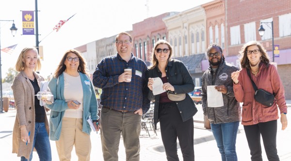 Six adults smile and hold coffee, pastries, and notebooks while standing on a sunny Main Street sidewalk lined with historic buildings and lamppost banners that read "Nevada Cubs."