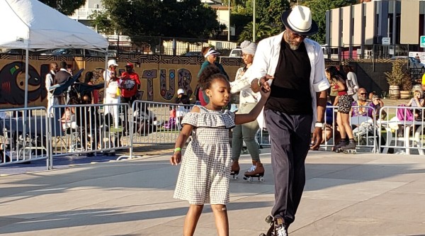 In East Oakland, California, a young girl and an older man hold hands while roller skating together at an outdoor community event hosted by the Black Cultural Zone.