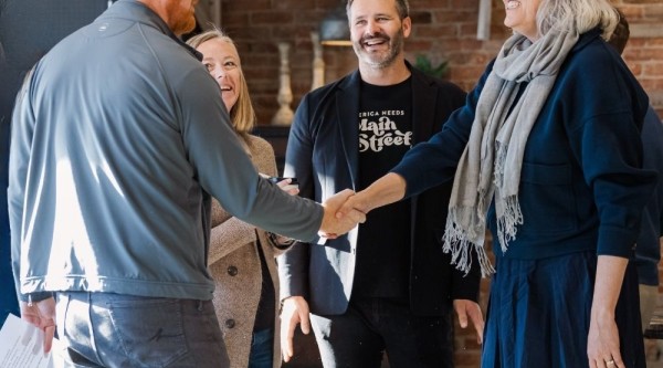 A group of five people greet each other inside a warmly lit coffee shop in Nevada, Iowa, with two individuals in the foreground shaking hands and others smiling in the background.