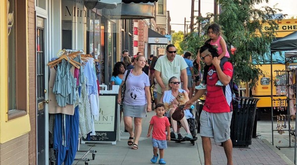 Shoppers stroll down a busy downtown sidewalk past a rolling rack of dresses and tops displayed outside a boutique. A child in a red shirt walks ahead of an adult pushing a stroller, while another adult carries a small child on their shoulders. Nearby, a person in a mobility scooter holds a small dog, and a street vendor’s jewelry stand and a yellow food truck peek out behind leafy trees.