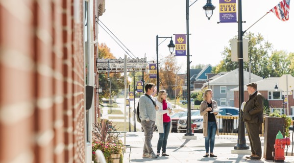 Four adults chat on a sunny downtown sidewalk in Nevada, Iowa, with purple “Nevada Cubs” banners and fall trees in the background.