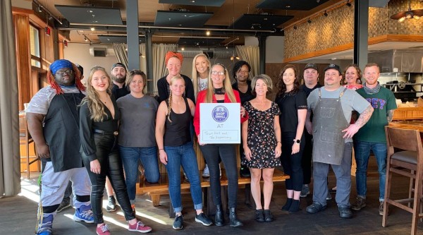 A group of about fifteen people stands together inside a modern restaurant space with wooden floors, high ceilings, and exposed ductwork. They wear a mix of work aprons, casual clothes, and business-casual outfits. In the center, one person holds a “Shop Small” sign that reads “AT Town Hall and The Dispensary.” Behind them are seating benches, tables, and an open kitchen area with tile accents on the walls.