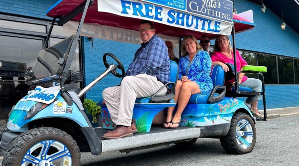 A custom-painted, tropical-themed golf cart parked on the street in front of a bright blue storefront, with a “FREE SHUTTLE” sign and sponsor banners overhead. Four passengers sit onboard—one at the wheel wearing a plaid shirt and light pants, two seated behind on blue cushions (one in a bright blue patterned dress), and one in a pink top—smiling at the camera. The cart’s body features palm tree and beach graphics, and its oversized off-road wheels are painted blue and white.