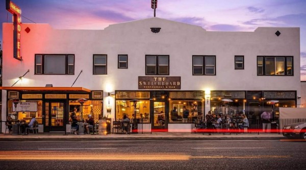 A two-story white stucco building at dusk, its ground floor glowing with warm light from large windows and outdoor seating areas on either side of a central entrance. A bronze sign above the door reads “The Switchboard Restaurant & Bar.” To the left, a neon vertical “Hotel” sign casts a red glow on the upper façade, while string lights and patio heaters illuminate diners outside. Faint streaks of red tail lights blur across the street in front, and a purple-pink sky fades into deep blue above.