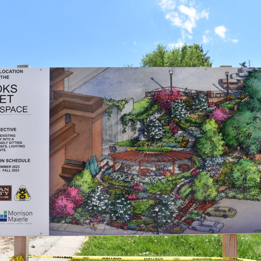 A construction site sign showing the future location of the Brooks Street Greenspace in Sheridan, Wyoming. The sign includes a colorful illustrated rendering of the planned park, featuring pathways, lush landscaping, a pergola seating area, and lighting. Text on the sign outlines the project objective and construction schedule, with logos from local partners and planning organizations.