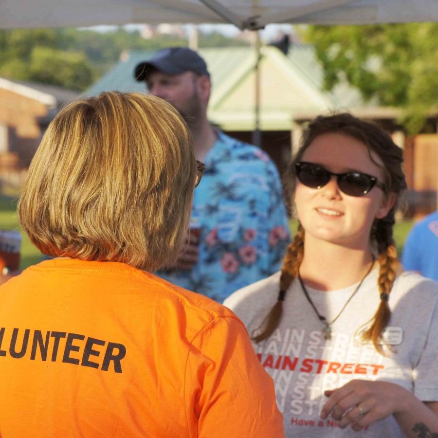 Volunteer in an orange shirt speaks with a Main Street staff member during an outdoor event, with others gathered casually in the background.