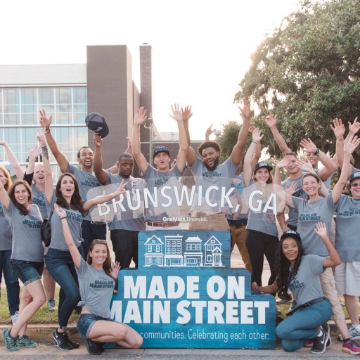 People cheer and smile while gathered around a sign that reads: "Brunswick, GA, Made on Main Street."