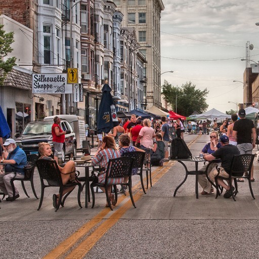 People gather around bistro tables in the middle of a street lined with historic buildings.