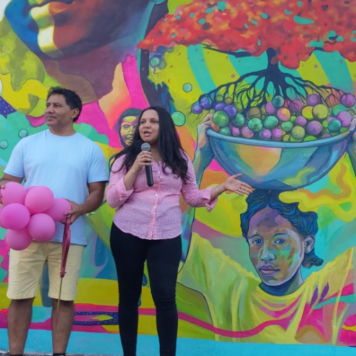 Two men and one woman stand in front of a brightly colored mural. The woman speaks into a microphone; to her right a man holds a cluster of balloons and a large pair of ceremonial scissors; the other man stands to her left.