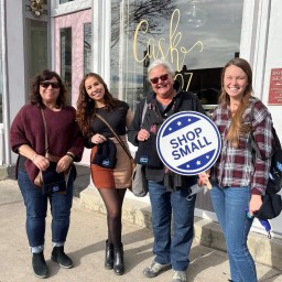 Four individuals stand shoulder-to-shoulder on a sunny sidewalk in front of a white-trimmed storefront with a pink door and gold script reading “Cask 27.” From left to right: one wearing a burgundy sweater and jeans, one in a black top and color-block skirt with tights and ankle boots, one in a black jacket and jeans holding a round blue-and-white “Shop Small” sign, and one in a plaid shirt and jeans. Each holds a black beanie featuring a local logo.