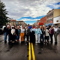 About twenty people stand shoulder-to-shoulder across the double yellow lines in the middle of a wet downtown street, smiling at the camera. They wear a mix of jackets, sweaters, jeans, and colorful pants, with historic brick storefronts, parked cars, and a cloudy-turned-blue sky overhead.