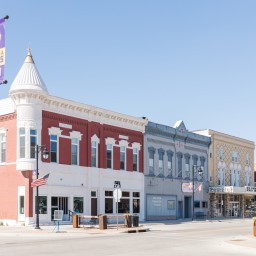Downtown Nevada, Iowa, featuring a row of historic brick buildings with colorful facades, including red, blue, and tan storefronts. American flags and a 'Nevada Cubs' banner hang from streetlamps, and outdoor seating is visible in the foreground under a clear blue sky.