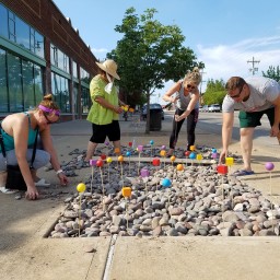 A group of four people works on a sidewalk art installation set within a recessed bed of river rocks. Dozens of slender wooden stakes topped with painted wooden shapes (spheres and cubes in bright yellow, orange, red, blue, and purple) rise from the stones. On the left, a crouching woman in a teal tank top and gray leggings picks up rocks; next to her, a standing woman in a wide-brimmed straw hat and green shirt holds two yellow-and-orange blocks. On the right, a woman in a gray tank top drives a stake into the stones with a mallet, while a man in a gray t-shirt and green shorts bends over to adjust a red cube on its stake. Behind them, a row of brick storefronts and the street with parked cars frame the scene under a clear blue sky.