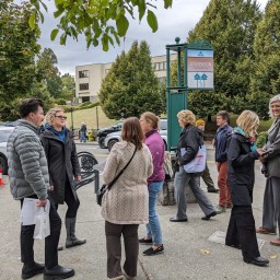 A group of people gathers on a sidewalk under leafy trees in downtown Stevenson, Washington. They appear to be engaged in conversation or a walking tour. A signpost labeled 'Stevenson, Washington' is visible in the background, along with parked vehicles and a tree-lined street.