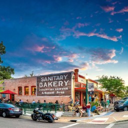 A vibrant streetscape at dusk showing a low brick building with a large painted sign that reads “SANITARY BAKERY – QUALITY AND SERVICE A. Ramsell, Prop.” Red-trimmed windows and a small “PIZZERIA” blade sign flank its corner entrance. In front, people sit at outdoor tables under red and green umbrellas, while others chat on a raised patio platform. A black motorcycle and several cars line the street beside a raised curb and crosswalk. Lush trees frame the scene under a deepening blue sky streaked with pink-tinged clouds.