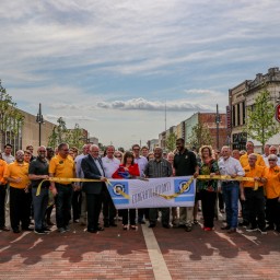 A large group of people gathered in downtown Denison, Texas, for a ribbon-cutting ceremony. They are holding a banner that reads 'Congratulations!' while standing on a newly renovated brick-paved street. The group includes community leaders, local officials, and residents, with a mix of yellow shirts, business attire, and casual wear. The setting is framed by restored historic buildings, trees, and a stop sign marking Main Street and Rusk Avenue.
