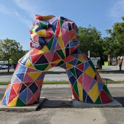 A large public sculpture of two oversized legs rising from square concrete bases and meeting at the top in a firm handshake. The entire piece is painted in a vibrant mosaic of geometric shapes—triangles and quadrilaterals in red, yellow, green, blue, pink, and purple—outlined in white. It stands on a roadside sidewalk with a parking lot, trees, and low buildings visible in the background.