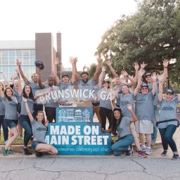 A group of about twenty people wearing matching “Made on Main Street” t-shirts stand and kneel behind a layered sign reading “Brunswick, GA” over “Made on Main Street – communities. Celebrating each other.” They’re outdoors on a street curb with event tents and a modern building behind them, and nearly everyone has arms raised in an enthusiastic gesture.