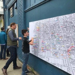 People in Covington, Kentucky add color-coded stickers to a large illustrated map mounted on a downtown building wall as part of a community engagement activity.