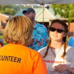 A person wearing a bright orange shirt with “VOLUNTEER” printed in bold on the back talks to someone facing the camera who is wearing sunglasses and a gray “Main Street” t-shirt; they stand under a canopy at an outdoor event with other attendees mingling in the background.