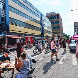 A sunny city street closed to traffic hosts a lively community event. In the foreground, three people sit at a small round table eating from take-out containers while another person at right films the scene with a DSLR camera. Behind them, attendees stand in line at a red “Award Winning BBQ” food truck, and pedestrians and bicyclists wander past art-deco–style and brick commercial buildings under a partly cloudy sky.