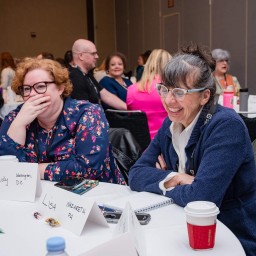 Two women seated at a round conference table share a laugh as they listen to a session. A red coffee cup and name cards sit in front of them, and other attendees are visible chatting at tables in the background.