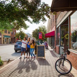 Three people stand on a brick-paved sidewalk beneath leafy tree branches, holding up a phone together to take a selfie. To their right, a sandwich-board sign advertises specials in front of a red-awning storefront, and a bicycle leans against the building. Parked cars line the street, and a parent pushes a stroller farther down the sidewalk under a banner–draped lamppost.