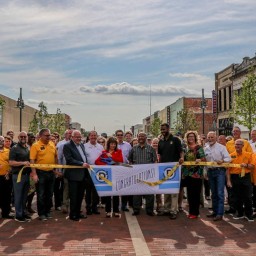 A large group of people stands across a freshly brick-paved street in front of new streetlights and storefronts. In the center, two individuals hold oversized ceremonial scissors poised over a yellow ribbon printed with “Congratulations!” as others grasp the ribbon at either end. Many in the crowd wear matching gold shirts, while others are dressed in business or casual attire. Young trees line the street, and a stop sign and “Main St” street sign are visible on the right.