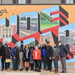 A group of about a dozen people in mixed business and casual attire stand side by side in front of a colorful brick-wall mural that reads “Greetings from Hyde Park,” with each letter filled by painted scenes of local landmarks. Most are wearing face masks, and a house’s siding is visible above the mural.