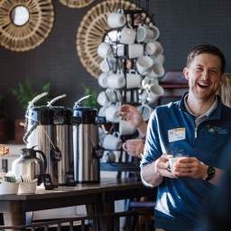 A smiling man wearing a blue Missouri Main Street Connection pullover and a name badge stands near a coffee station, holding a steaming white mug. Behind him, a rotating mug rack and wicker baskets of stirrers and condiments sit atop the counter, while decorative woven wall mirrors and natural light fill the cozy café setting.