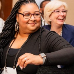 A conference attendee with long braided hair, wearing glasses, hoop earrings, and a black top, sits at a table smiling and looking to her left. She has a name badge around her neck and a smartwatch on her wrist. Behind her, another attendee with short blonde hair and glasses laughs, and a third person sits further back in a patterned jacket.