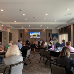 A small conference room set up with round tables where about a dozen participants sit facing the front. At the head of the room, five panelists are seated in front of a large wall‐mounted screen displaying “Downtown Florence,” and a masked presenter stands beside them holding notes. Attendees around the tables are reviewing papers, sipping water, and listening—some wearing masks—and laptops, notebooks, and beverage bottles are spread across the tables.