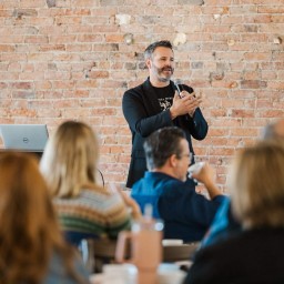 A man stands at the front of a room holding a microphone and gesturing as he speaks. He wears a black blazer over a dark t-shirt and stands in front of an exposed brick wall. In the foreground, several seated attendees—seen from behind—listen, one sipping from a cup. A laptop sits on a small table beside the speaker.