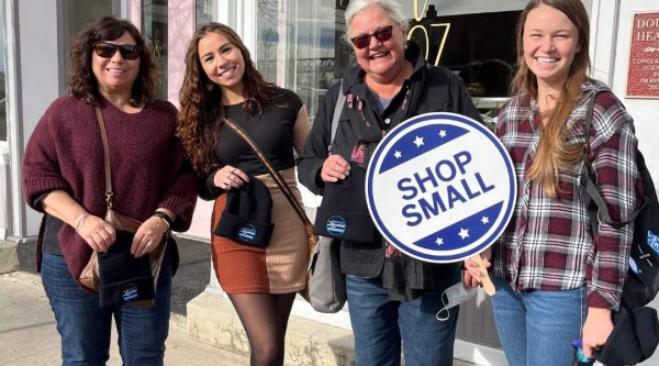 Four individuals stand shoulder-to-shoulder on a sunny sidewalk in front of a white-trimmed storefront with a pink door and gold script reading “Cask 27.” From left to right: one wearing a burgundy sweater and jeans, one in a black top and color-block skirt with tights and ankle boots, one in a black jacket and jeans holding a round blue-and-white “Shop Small” sign, and one in a plaid shirt and jeans. Each holds a black beanie featuring a local logo.