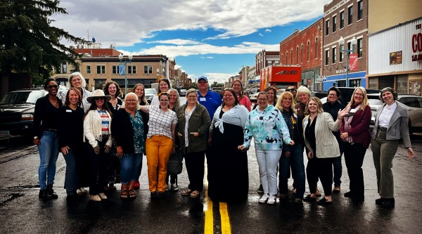 About twenty people stand shoulder-to-shoulder across the double yellow lines in the middle of a wet downtown street, smiling at the camera. They wear a mix of jackets, sweaters, jeans, and colorful pants, with historic brick storefronts, parked cars, and a cloudy-turned-blue sky overhead.