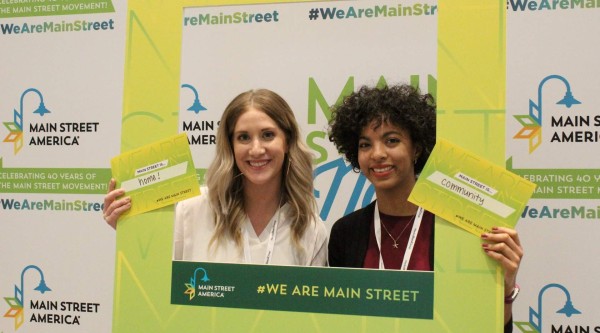 Two smiling women pose behind a large lime-green frame branded “Main Street America” and “#WeAreMainStreet.” Each holds a small card: the woman on the left’s reads “Main Street is… home!” and the woman on the right’s reads “Main Street is… community.” Behind them is a repeating backdrop celebrating 40 years of the Main Street movement.