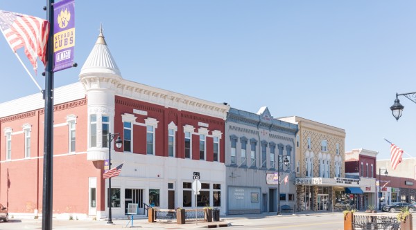 Downtown Nevada, Iowa, featuring a row of historic brick buildings with colorful facades, including red, blue, and tan storefronts. American flags and a 'Nevada Cubs' banner hang from streetlamps, and outdoor seating is visible in the foreground under a clear blue sky.