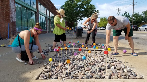 A group of four people works on a sidewalk art installation set within a recessed bed of river rocks. Dozens of slender wooden stakes topped with painted wooden shapes (spheres and cubes in bright yellow, orange, red, blue, and purple) rise from the stones. On the left, a crouching woman in a teal tank top and gray leggings picks up rocks; next to her, a standing woman in a wide-brimmed straw hat and green shirt holds two yellow-and-orange blocks. On the right, a woman in a gray tank top drives a stake into the stones with a mallet, while a man in a gray t-shirt and green shorts bends over to adjust a red cube on its stake. Behind them, a row of brick storefronts and the street with parked cars frame the scene under a clear blue sky.