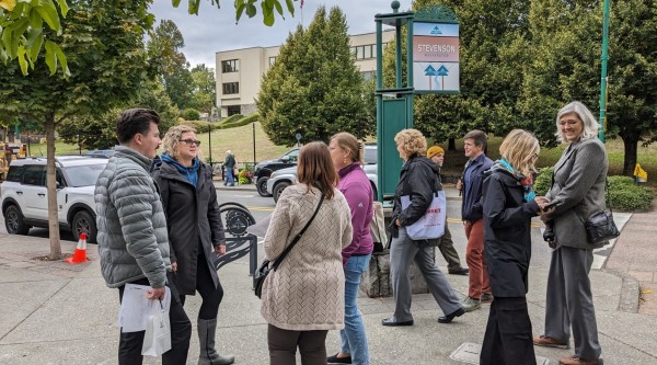 A group of people gathers on a sidewalk under leafy trees in downtown Stevenson, Washington. They appear to be engaged in conversation or a walking tour. A signpost labeled 'Stevenson, Washington' is visible in the background, along with parked vehicles and a tree-lined street.