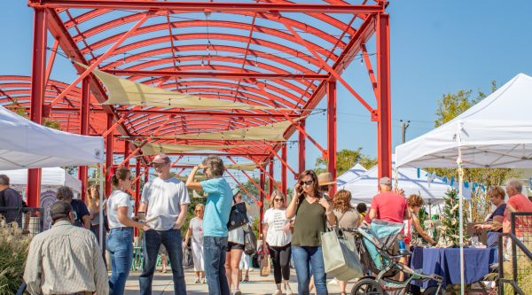 On a bright and sunny day, people shop at a farmers' market. A vibrant red archway spans the pavilion where vendors are set up under pop-up canopy tents.